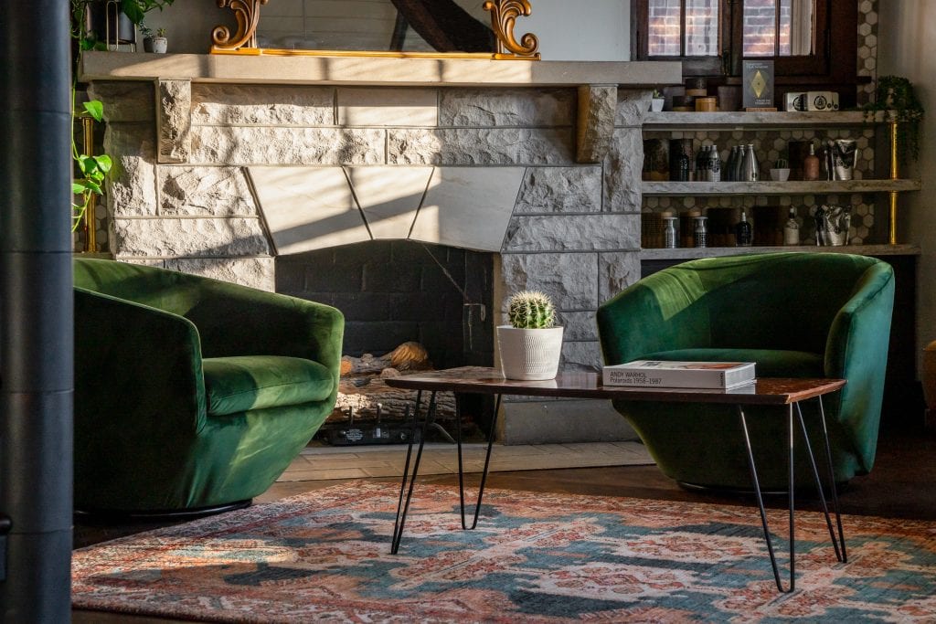 Bangarang 4 Green velvet chairs in a sunlit seating area with a stone fireplace and patterned rug.